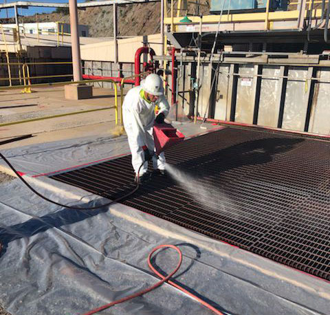 Man in white protective suit spraying gray primer onto rusted grate Maxon's CRS Coating prevents corrosion